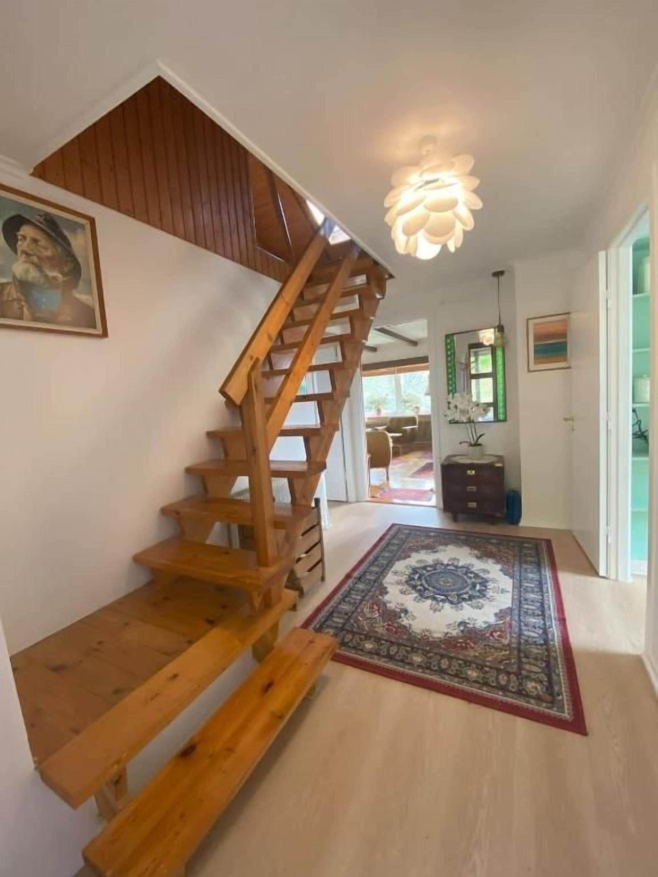 Una escalera de madera conduce a un dormitorio tipo loft, con un banco de madera, una alfombra estampada y una lámpara de techo.