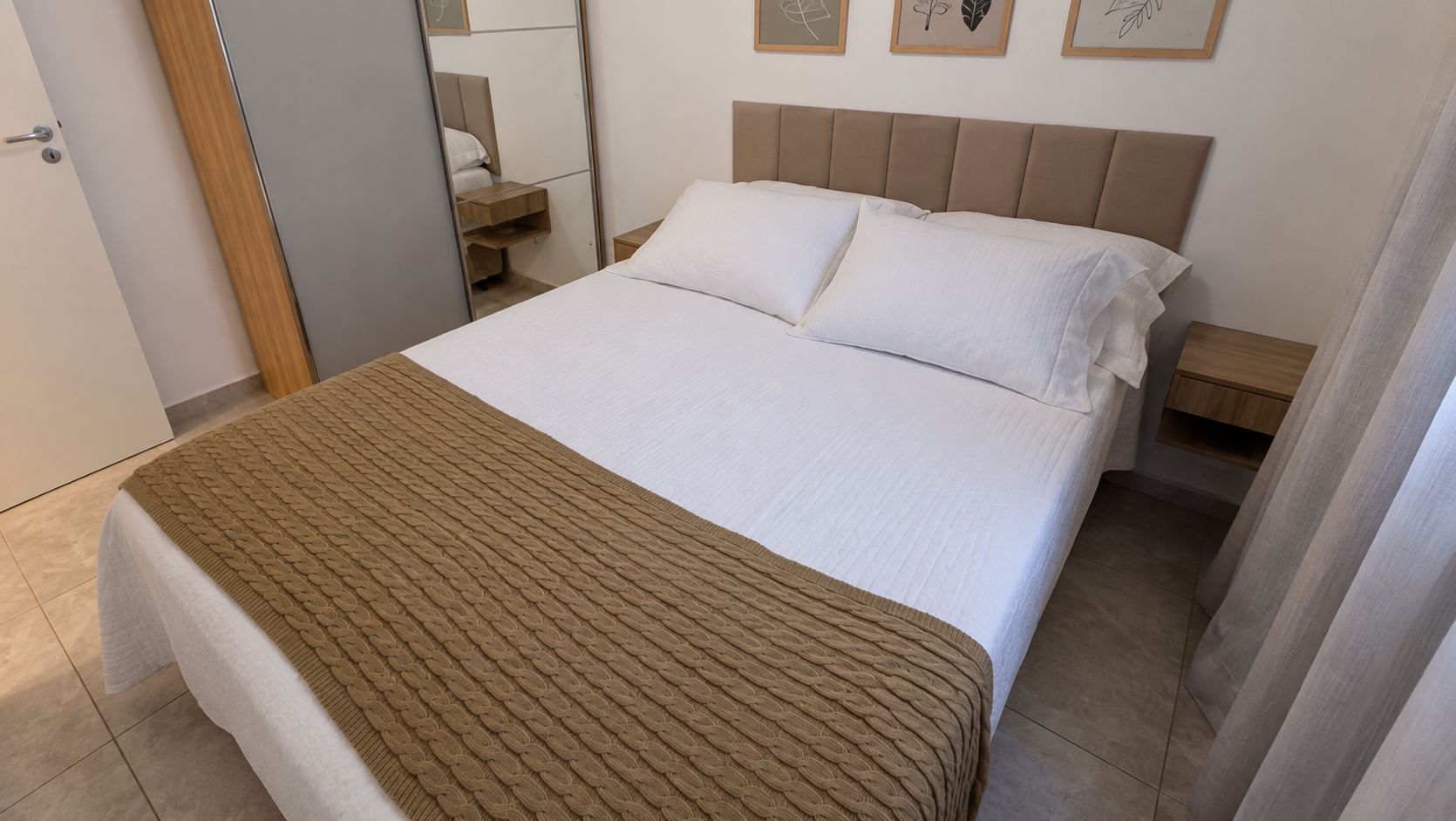 Ein ordentlich bezogenes Bett mit einer braunen Decke, ein Nachttisch mit einer Lampe und ein an der Wand befestigtes Regal.