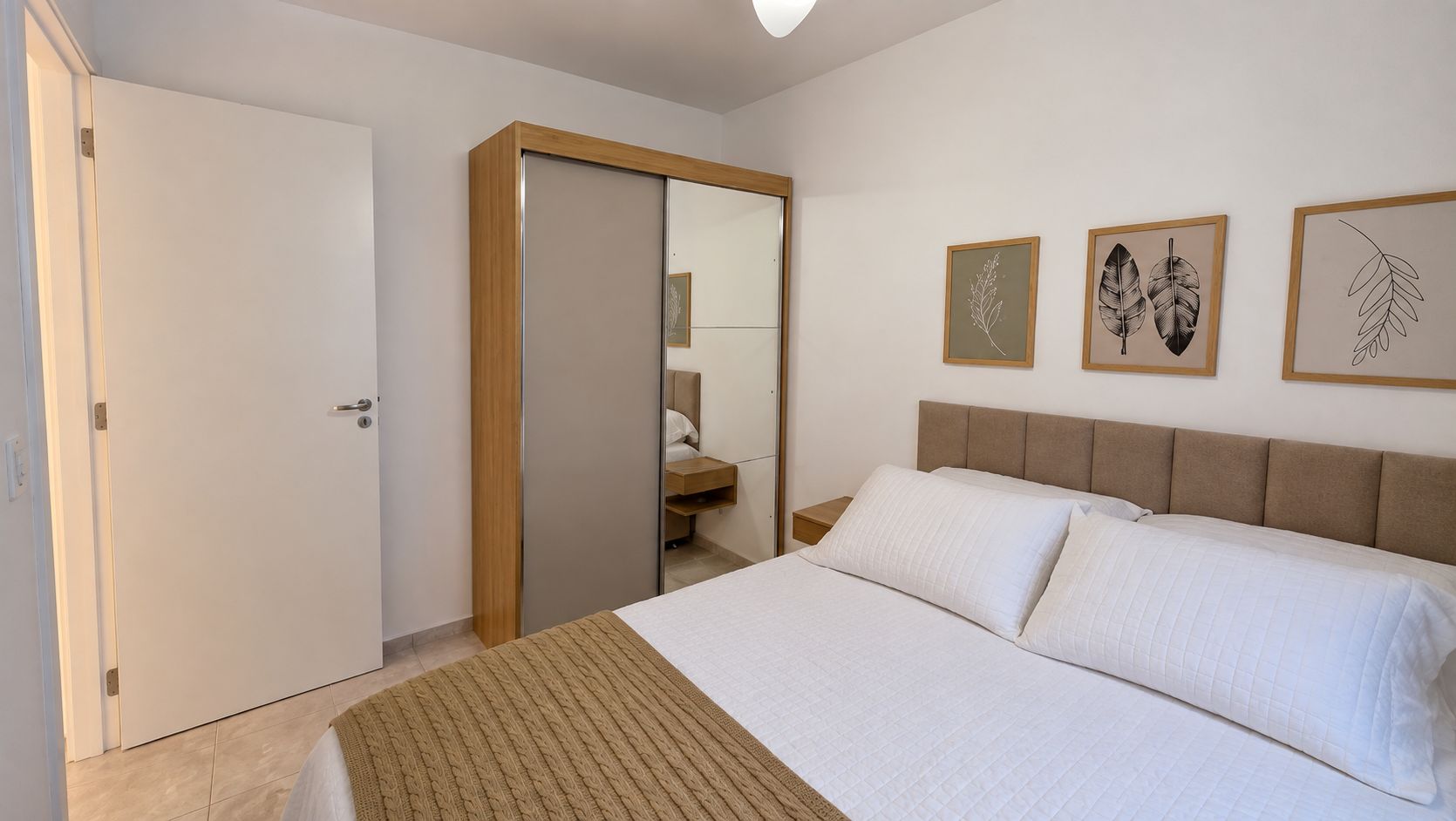 A bedroom with a bed, a wardrobe, a mirror, and framed botanical prints on the wall.