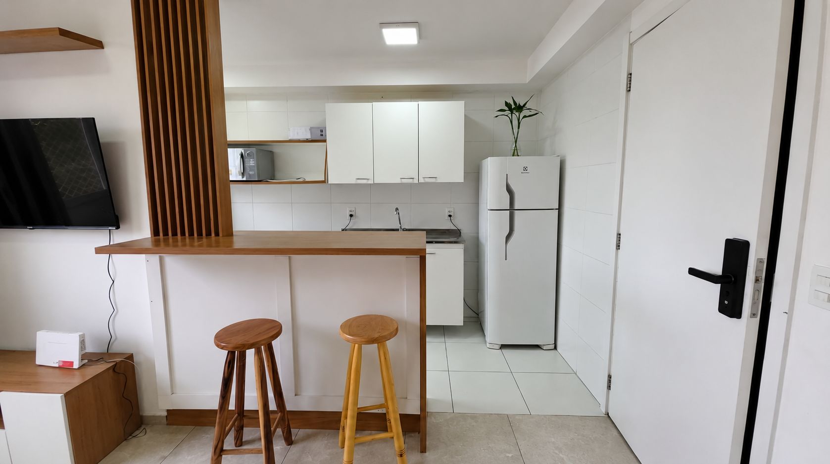 A compact kitchen with a wooden bar counter, two stools, a refrigerator, and a microwave.