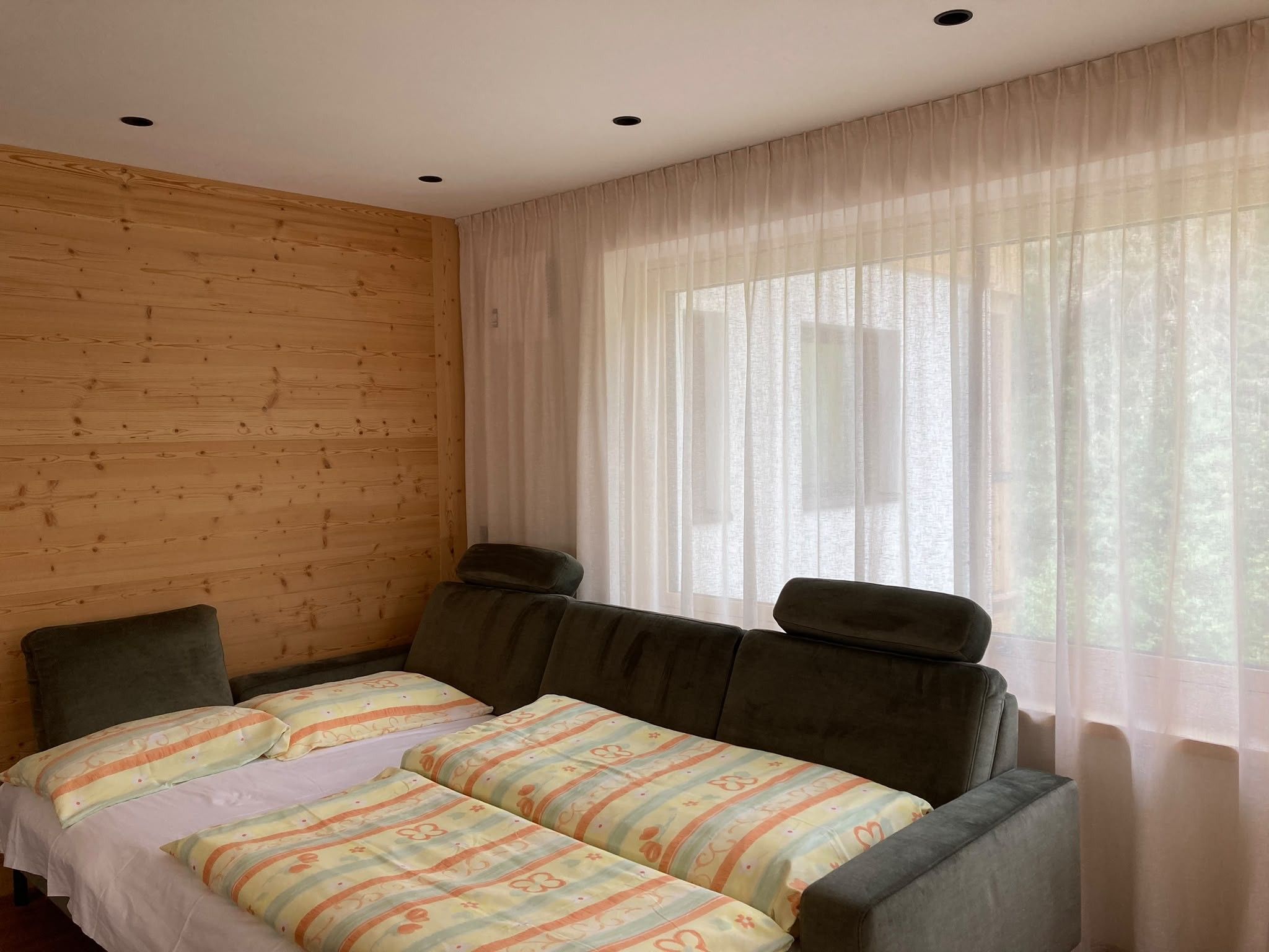 Ein Zimmer mit einer Holzwand, einem Bett mit gemusterten Textilien und durchsichtigen Vorhängen.