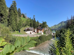 A chalet-style building with a river in the foreground, surrounded by dense forest and hills. - Apartment 'Pe De Munt Mez Col' with Mountain View, Balcony and Wi-Fi (La Valle)