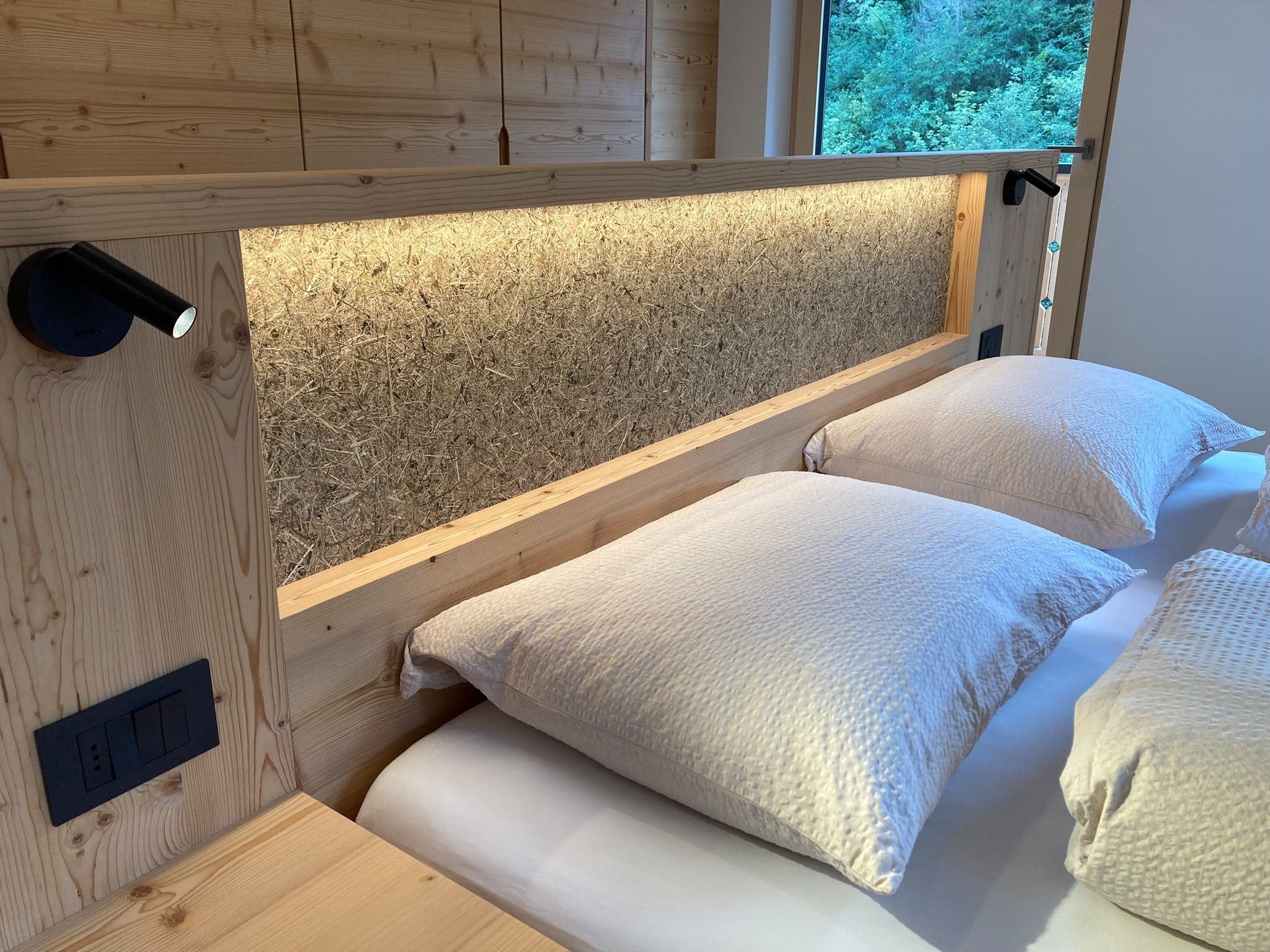 Una cama litera de madera con cabecera de listones y una ventana con vistas a la vegetación.
