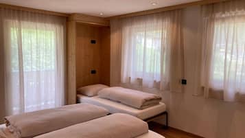 A room with two beds, wooden walls, and large windows with sheer curtains.