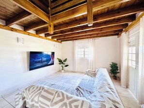 A room with a wooden ceiling, a flat-screen TV mounted on the wall, a sofa, a potted plant, and a window with curtains. - Villa Between Land and Sea (Les Anses-d'Arlet)