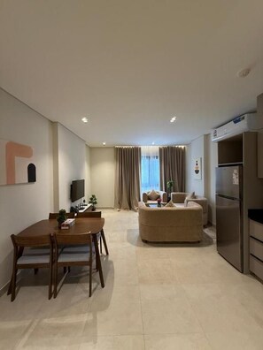 A modern living room with a sofa, a dining table with chairs, a flat-screen TV, and a kitchen area with a refrigerator.