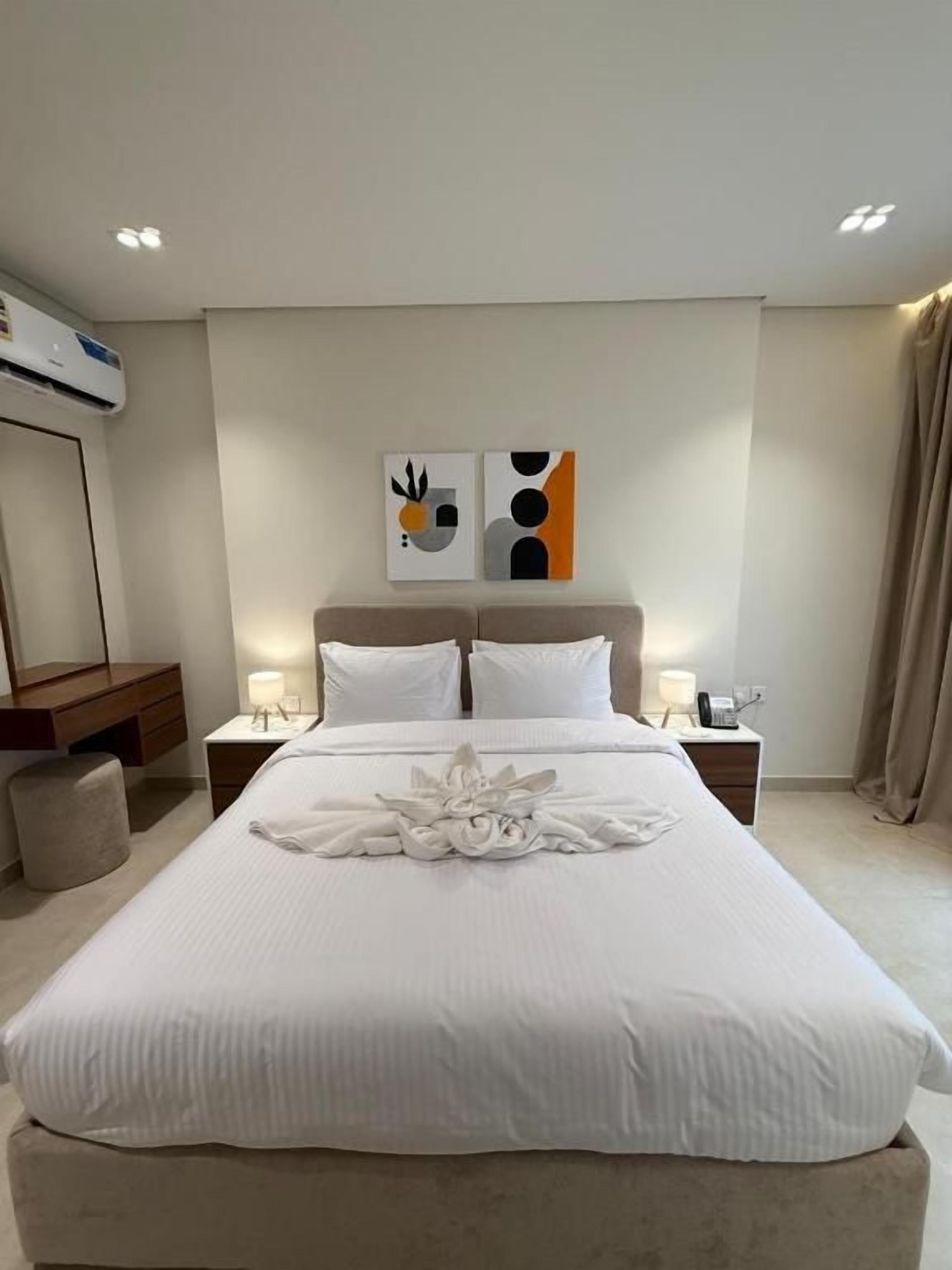 Une chambre d’hôtel avec un grand lit, une table de chevet avec une lampe, un climatiseur fixé au mur et une œuvre d’art abstraite au-dessus du lit.