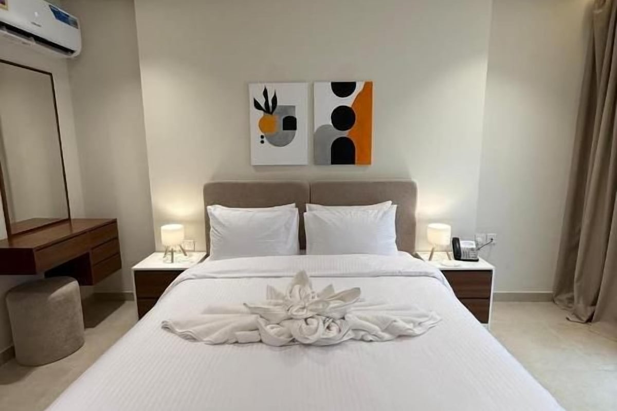 A hotel room with a large bed, a bedside table with a lamp, a wall-mounted air conditioner, and abstract artwork above the bed.