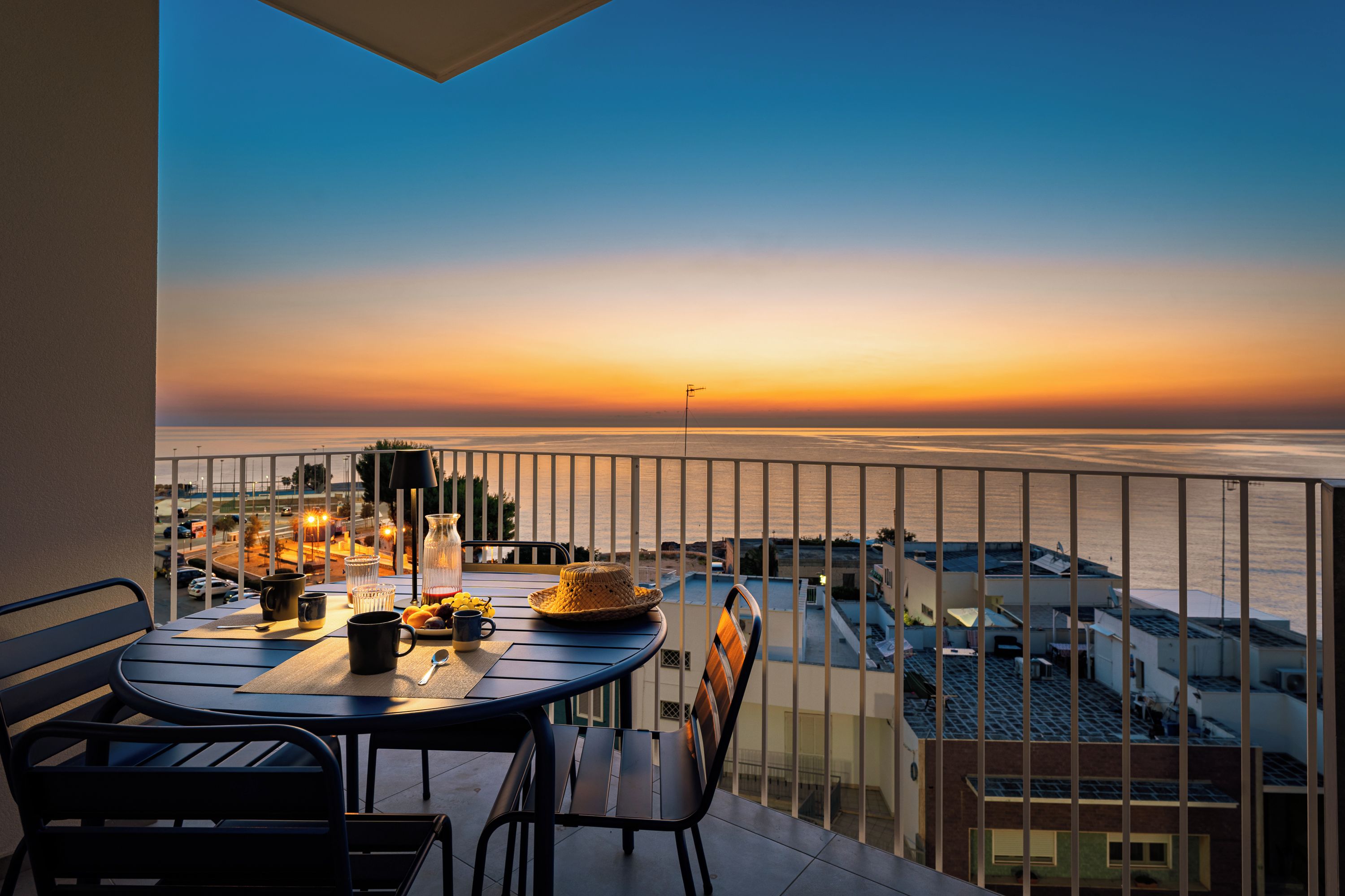 A balcony with a table set for two, overlooking a sunset over the sea.
