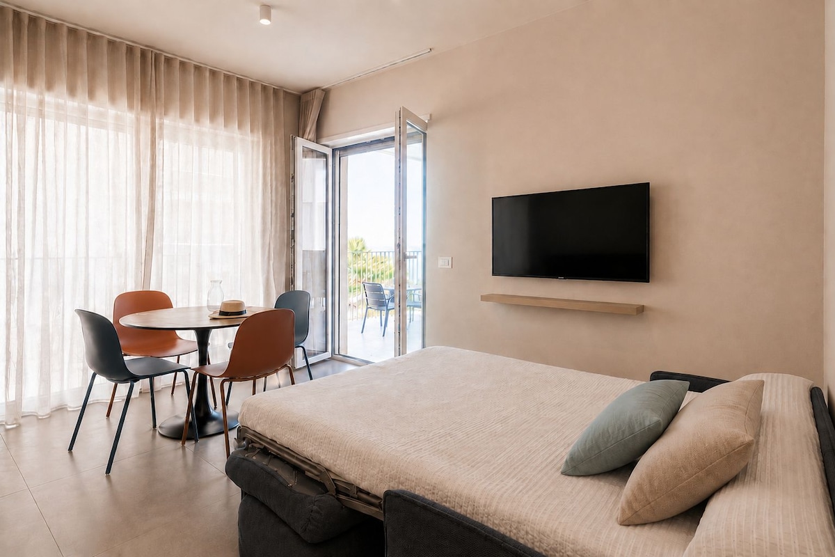 A hotel room with a bed, a TV mounted on the wall, a dining table with chairs, and a view through the window.
