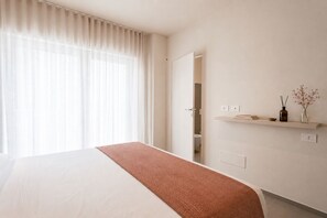 Habitación de hotel con cama, ventana con cortinas, puerta que da a un baño y estante con un florero y libros.