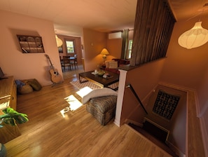 A spacious living area with a wooden floor, a sofa, a coffee table, and a guitar. A staircase with a patterned rug leads to a dining area. - Carmella Cottage, Antigonish (Antigonish)