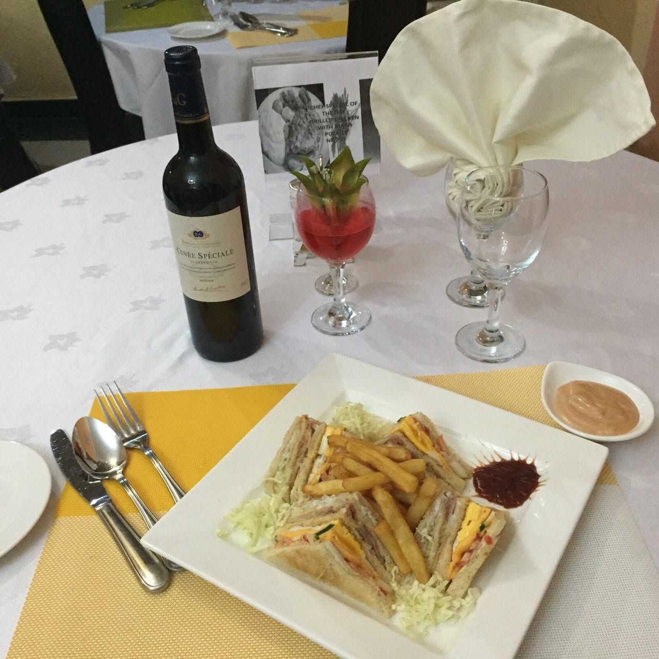 Une table dressée avec une bouteille de vin, un verre de vin rouge, un verre d’eau, une assiette de sandwichs, des frites et un petit bol de sauce.