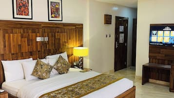 A hotel room with a wooden headboard, a large bed with white linens, a red rug, a TV on a wooden stand, and two framed artworks on the wall.