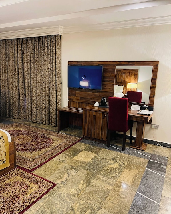 Habitación de hotel con un escritorio de madera, televisión, una silla roja, un espejo grande y una alfombra estampada.