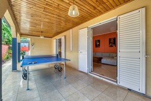 An indoor space with a ping pong table, a ceiling fan, and a door leading to a living area with a sofa and artwork. - Villa Galéa - Climatisée, vue mer, piscine (Etang-Salé les Hauts)