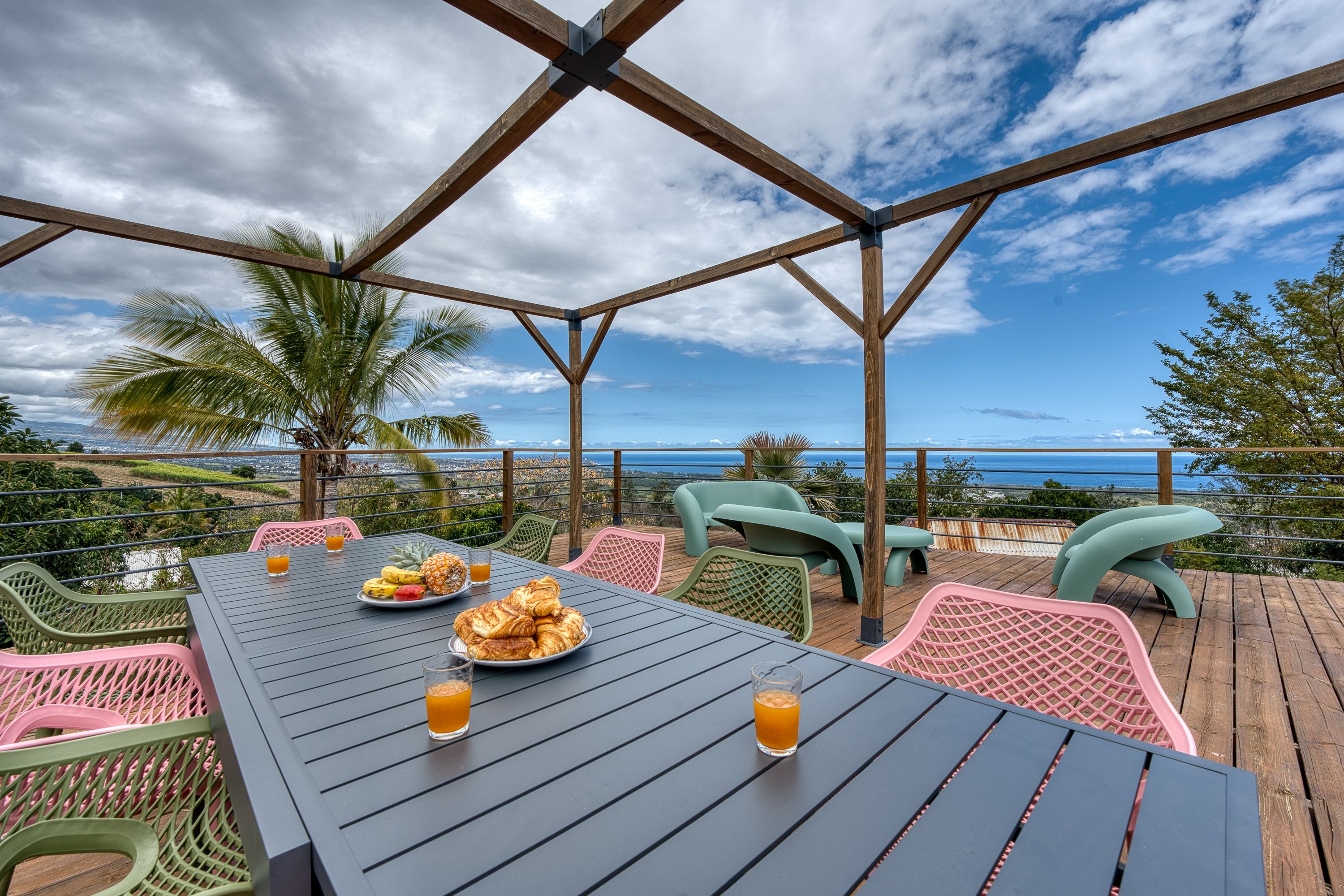 Un espace de restauration en plein air offrant une vue sur l’océan, avec une terrasse en bois, des chaises à structure métallique et une table dressée avec de la nourriture et des boissons.