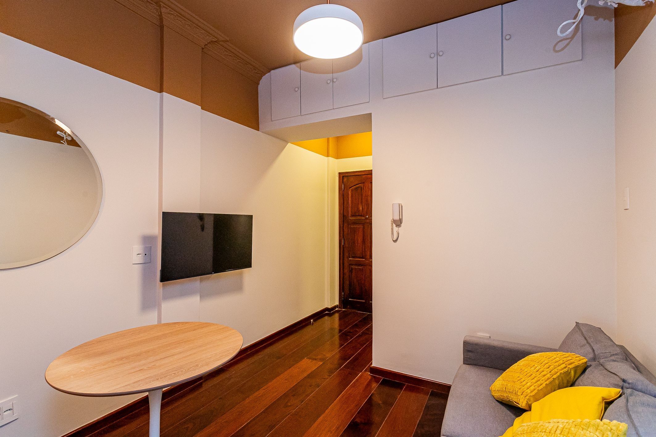 A compact living space with a wooden floor, a round table, a sofa with yellow cushions, a mounted TV, and a door leading to another room.