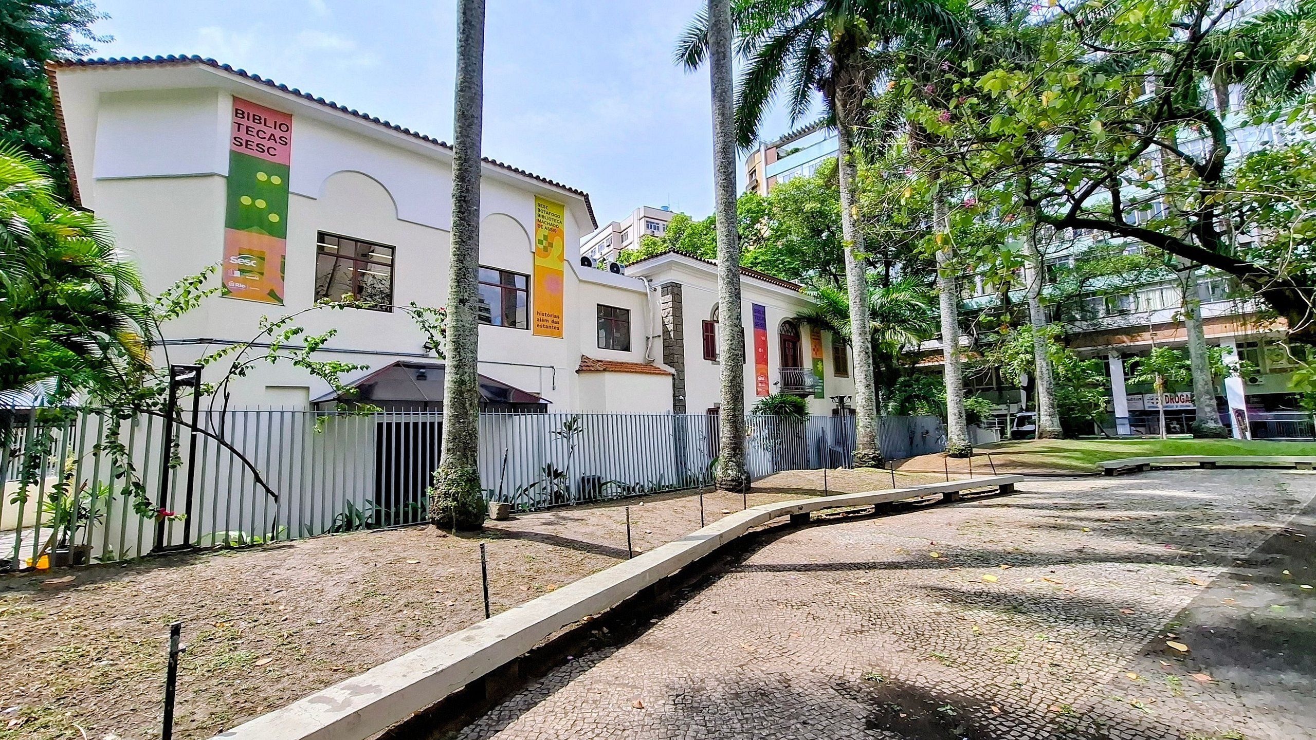 A building with multiple colorful banners, a paved pathway, and a fenced area with trees.