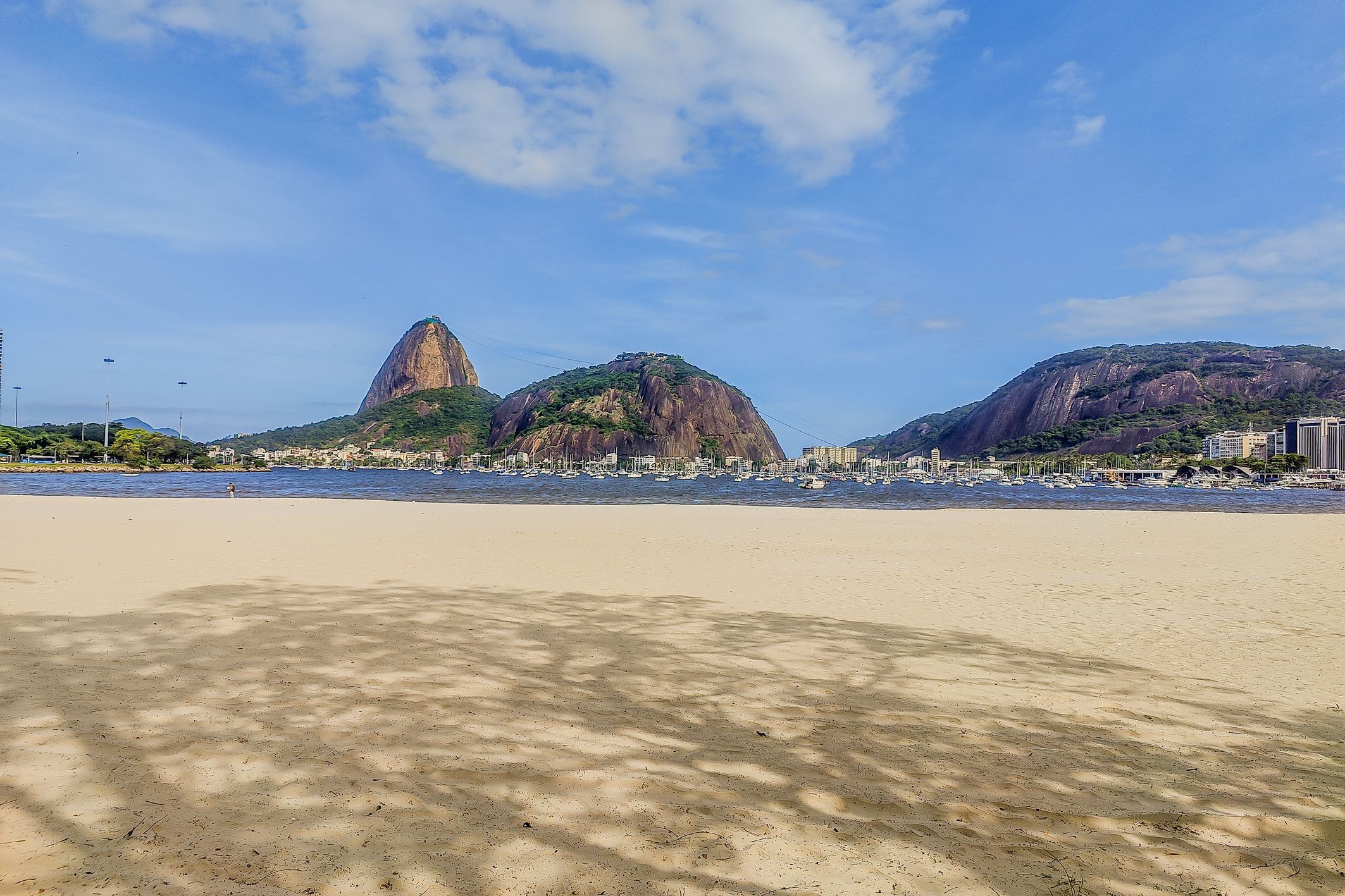 A beach with a prominent rock formation in the background, a clear sky, and a calm sea with boats.