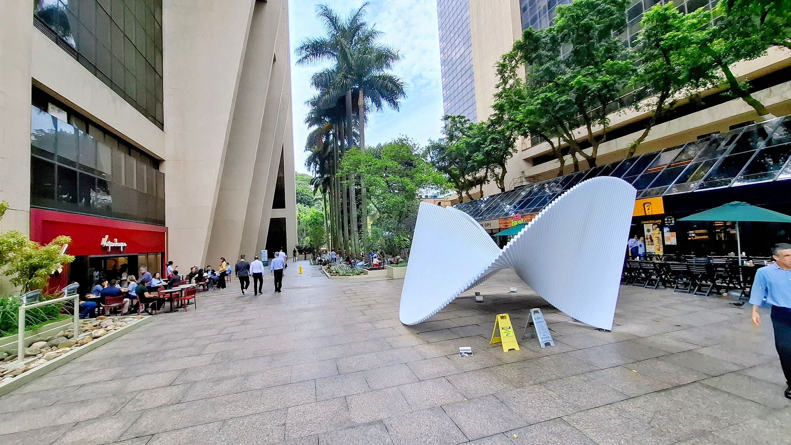 An urban plaza with a modern sculpture, cafes, and a restaurant.