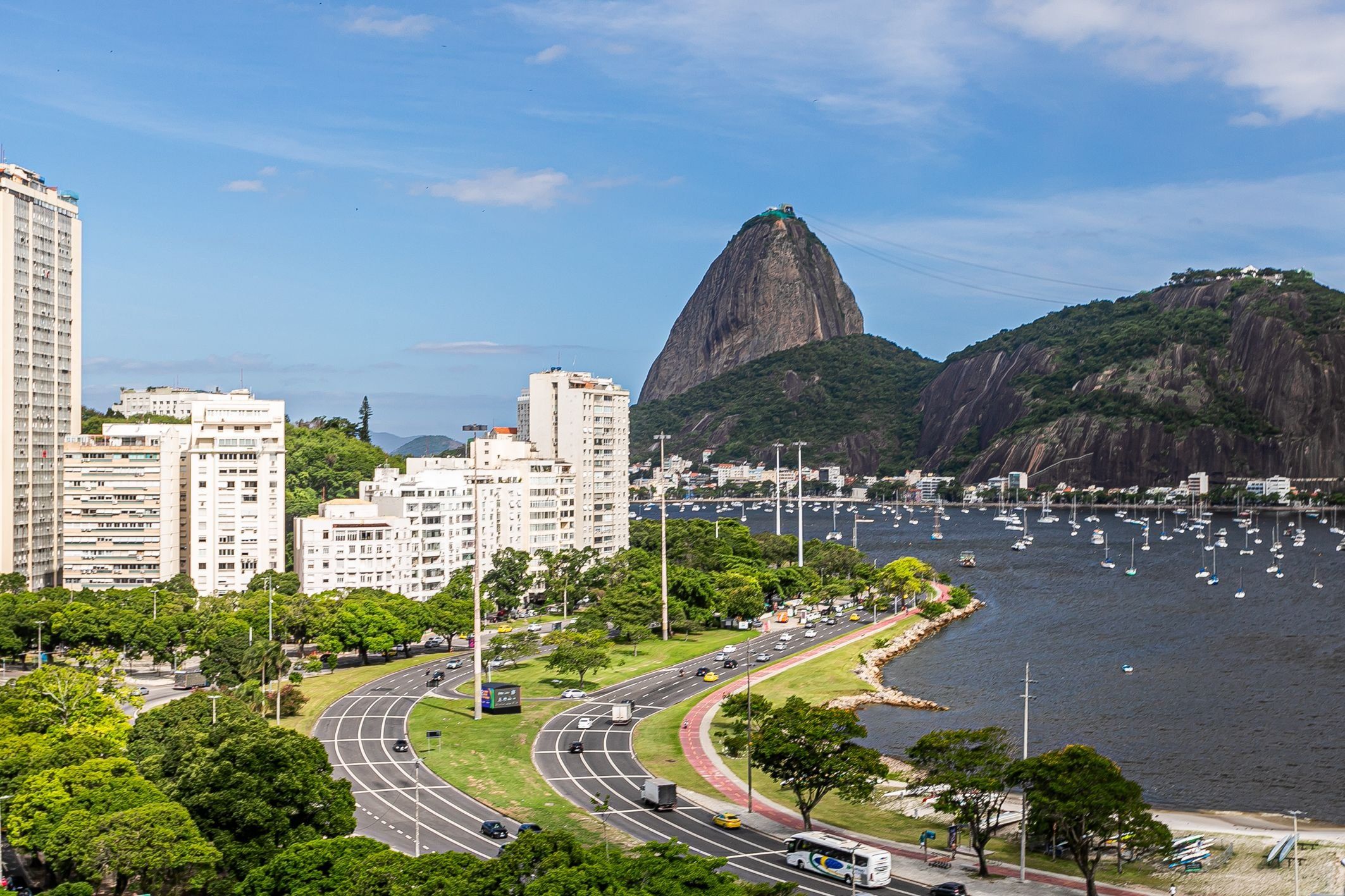 A coastal cityscape with a prominent mountain, a marina with boats, and a curved road with vehicles.