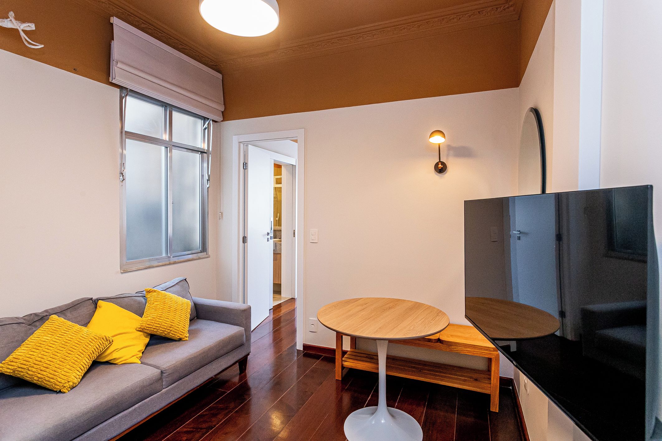 A modern living room with a sofa, a round wooden table, a flat-screen TV, and a window with blinds.