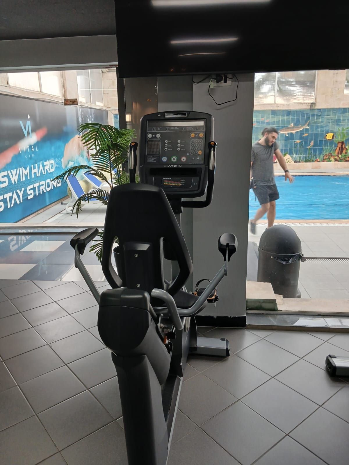 Un gimnasio interior con equipo de ejercicio, vista a una piscina y un cartel en la pared.