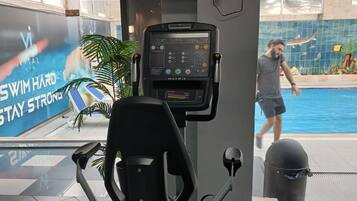 Un gimnasio interior con equipo de ejercicio, vista a una piscina y un cartel en la pared.