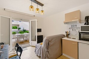 A compact kitchenette with a microwave, a small refrigerator, and a sink. There is a dining area with a table and chairs, and a covered sofa. The room has a sliding glass door leading to an outdoor area with a view of greenery. - À Deux Pas de Pentrez - Proche de la Plage (Saint-Nic)