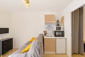 Ein kompakter Wohnbereich mit einer Küchenzeile, einem Bett mit gelbem Kissen, einer schwarzen Kommode und einem Fernseher.
