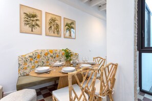Een eetgedeelte met een houten tafel voor vier personen, omringd door rieten stoelen, een bankje en een bank met een bloemenprint.
