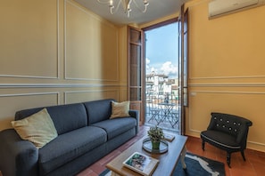 A room with a sofa, a chair, a coffee table with a book and a plant, and a view of buildings through an open door. - Sunflower Suite by MMega (Firenze)