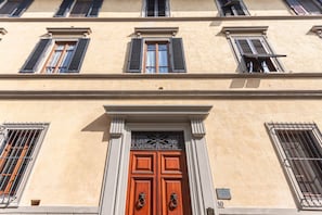 A building facade with a central entrance featuring a double wooden door with metal handles, flanked by windows with shutters and a decorative metal grille above the door. - Sunflower Suite by MMega (Firenze)
