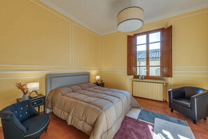 A bedroom with a large bed, a nightstand, a chair, a window with a view, and a radiator. - Sunflower Suite by MMega (Firenze)