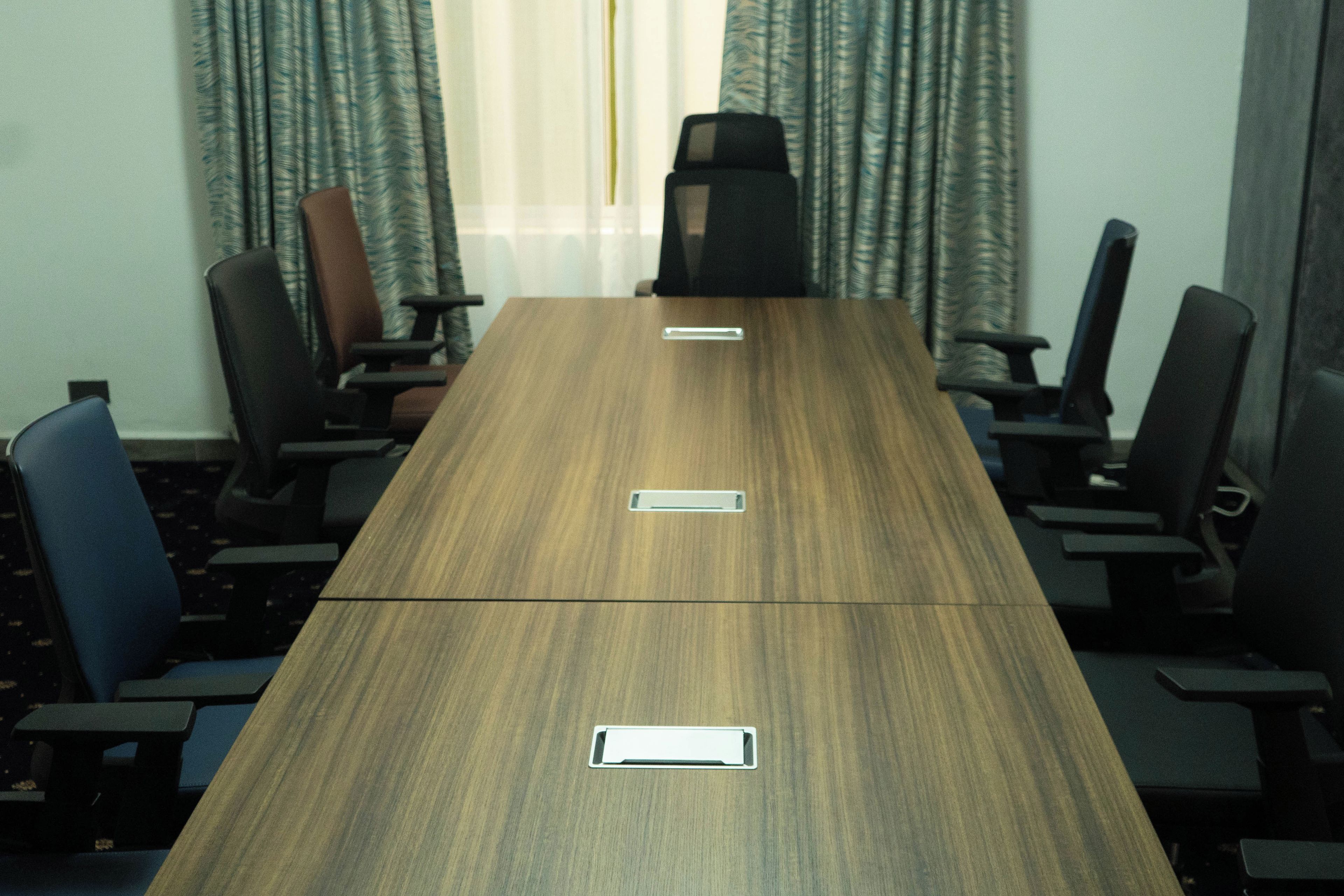 A conference room with a long wooden table, six chairs, and a window with curtains.