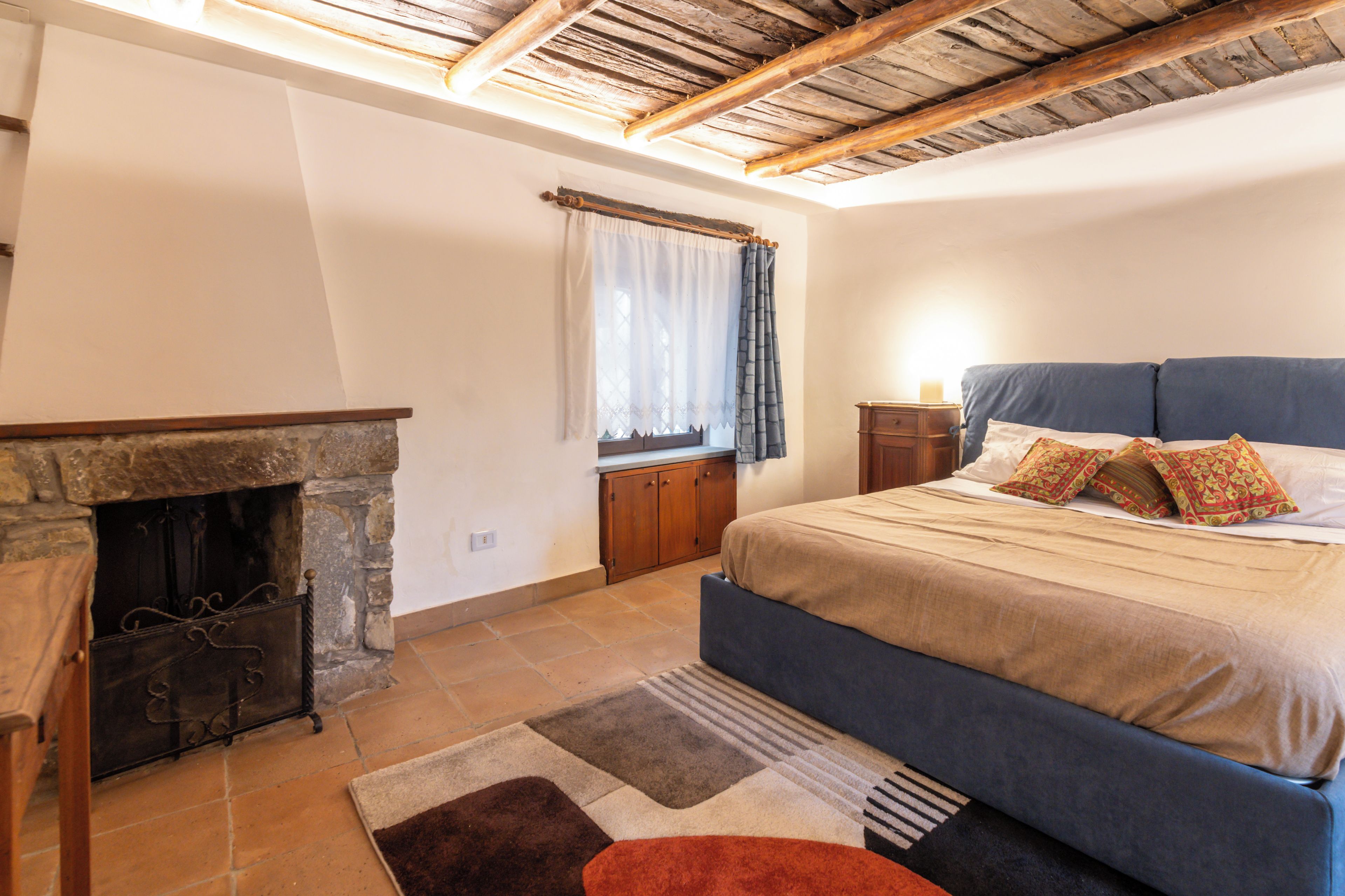 Ein Schlafzimmer mit Kamin aus Stein, einem Bett mit blauer Bettwäsche, einem Nachttisch und einem Fenster mit Vorhängen.
