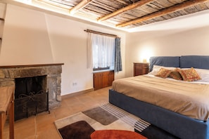 A bedroom with a stone fireplace, a bed with blue bedding, a nightstand, and a window with curtains. - Borgo 1660 - Dimora di Charme (Castellabate)