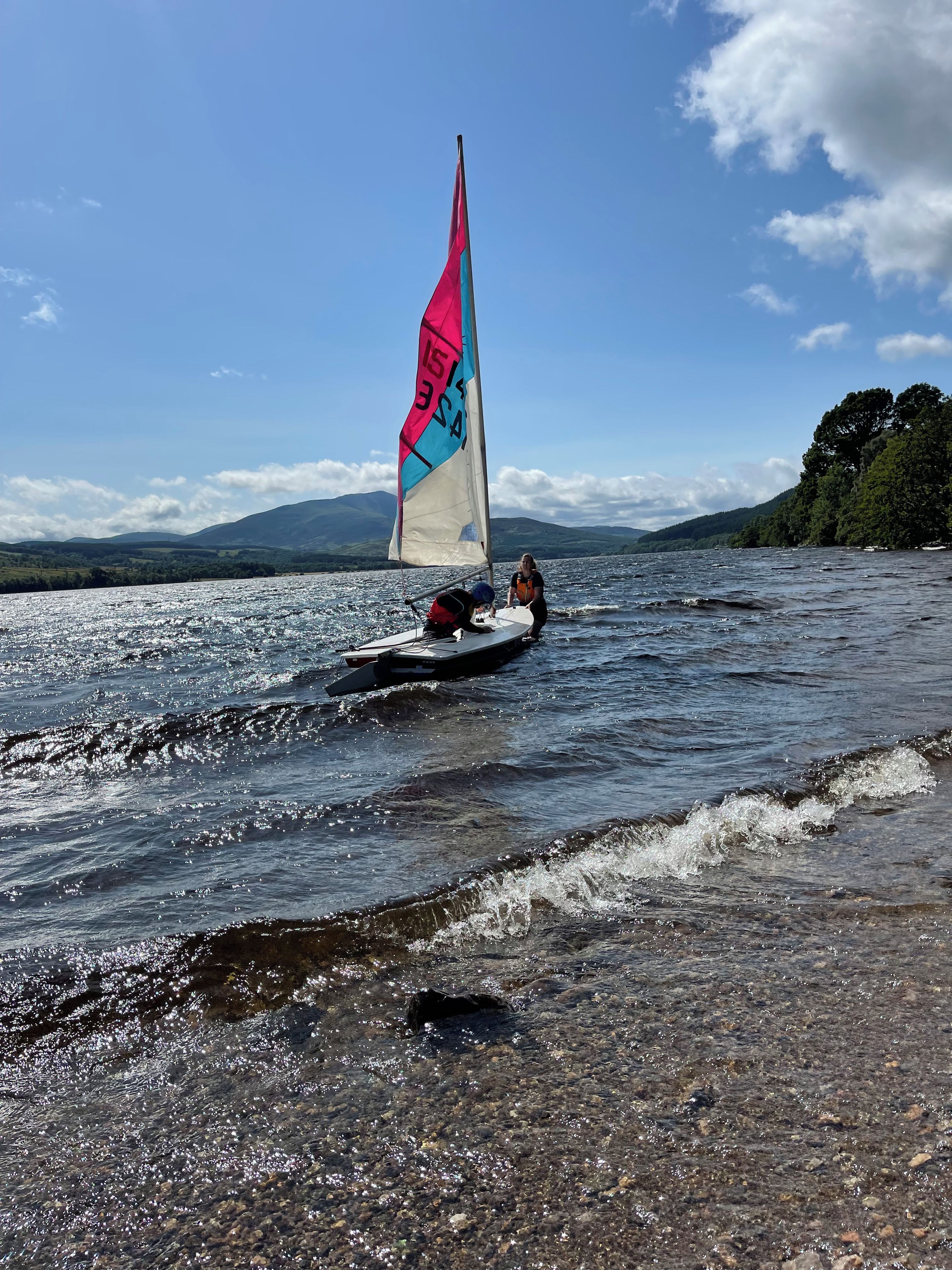 Un voilier sur un lac paisible, sous un ciel dégagé, avec des montagnes qui se dessinent à l’horizon.