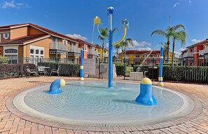 Une fontaine avec trois jets d’eau, entourée d’un chemin pavé de briques, avec des immeubles d’habitation en arrière-plan.
