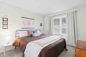 A bedroom with a bed, a bedside table, a lamp, a window with curtains, and wall decorations. - Lynnhaven Landing – Cozy Springfield Getaway (Springfield)