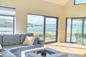 Ein modernes Wohnzimmer mit einem grauen Sofa, einem Couchtisch und Blick auf einen See und Hügel durch große Glasfenstertüren.