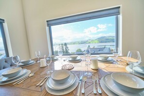 Une table à manger dressée avec des assiettes, des bols, des couverts et des verres, offrant une vue panoramique sur un lac et des collines, visible à travers une grande fenêtre.