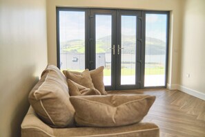 Une pièce avec un canapé beige, une porte-fenêtre coulissante donnant sur un balcon et une vue sur un paysage verdoyant.