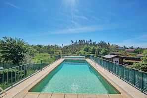 A rectangular swimming pool with a clear blue water surface, surrounded by a green fence, and set against a backdrop of trees and buildings. - Cozy vacation home surrounded by nature—relax and recharge (Leiria)