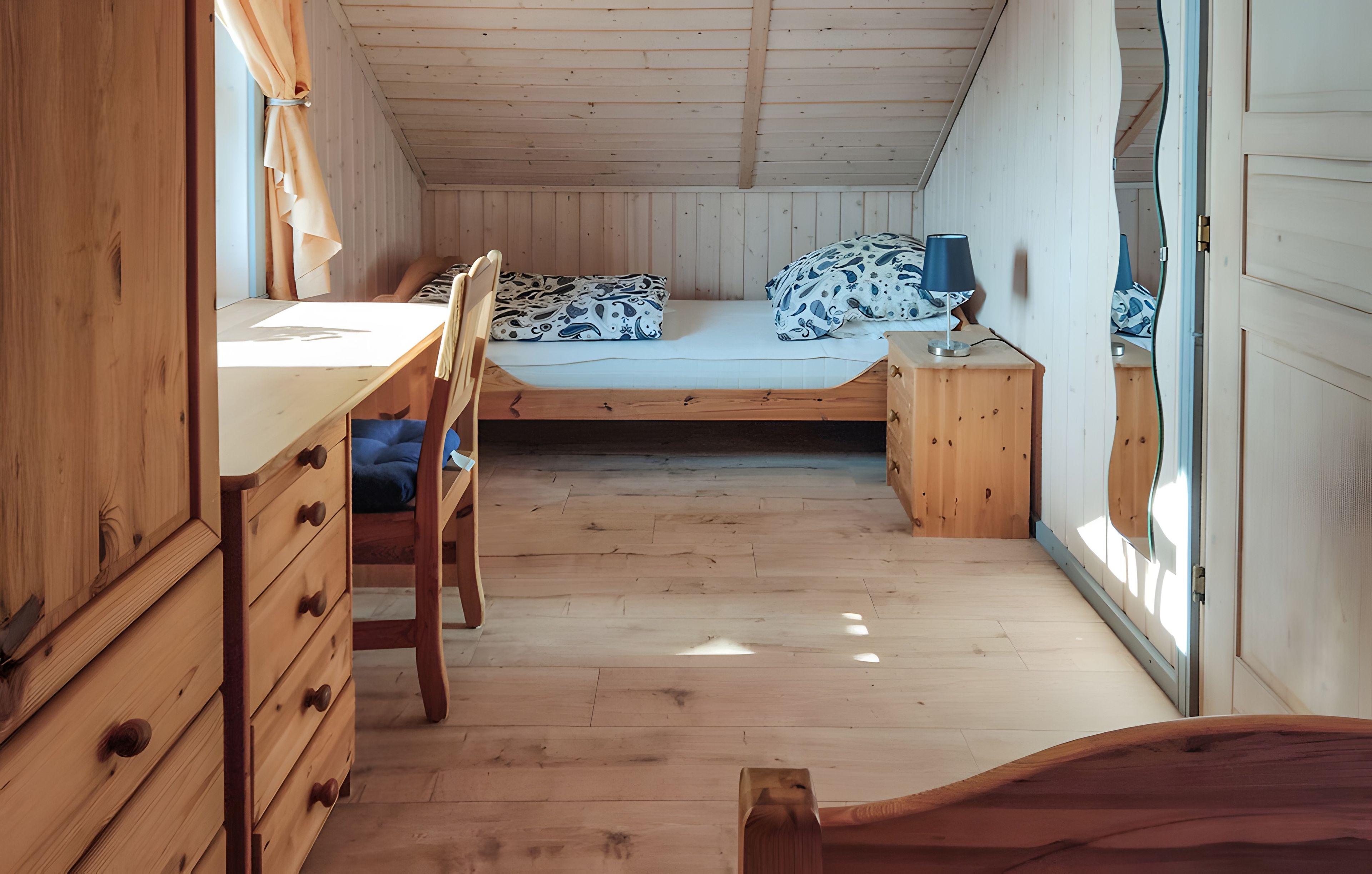 A compact room with a wooden desk, a single bed with patterned bedding, and a small bedside table with a lamp.