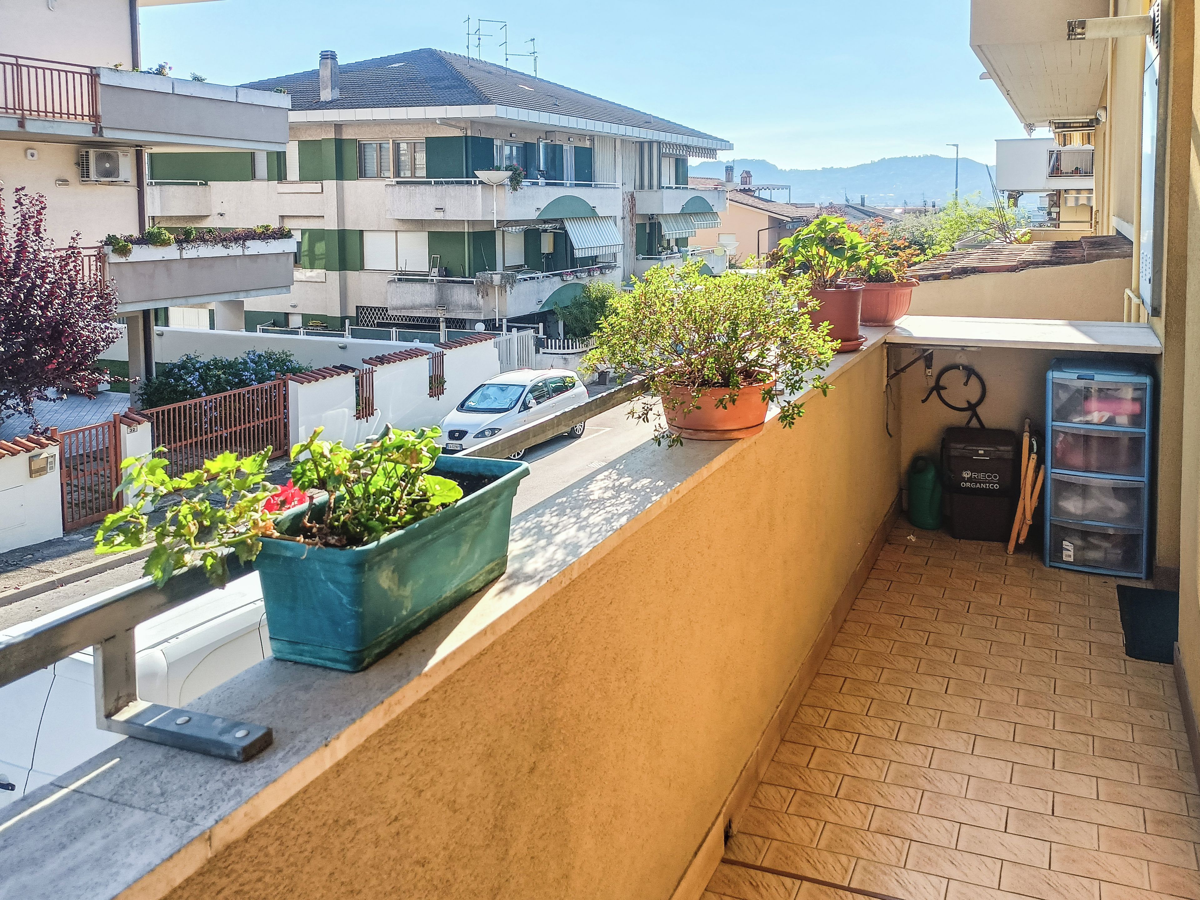 A balcony with potted plants, a trash bin, and a blue storage unit.