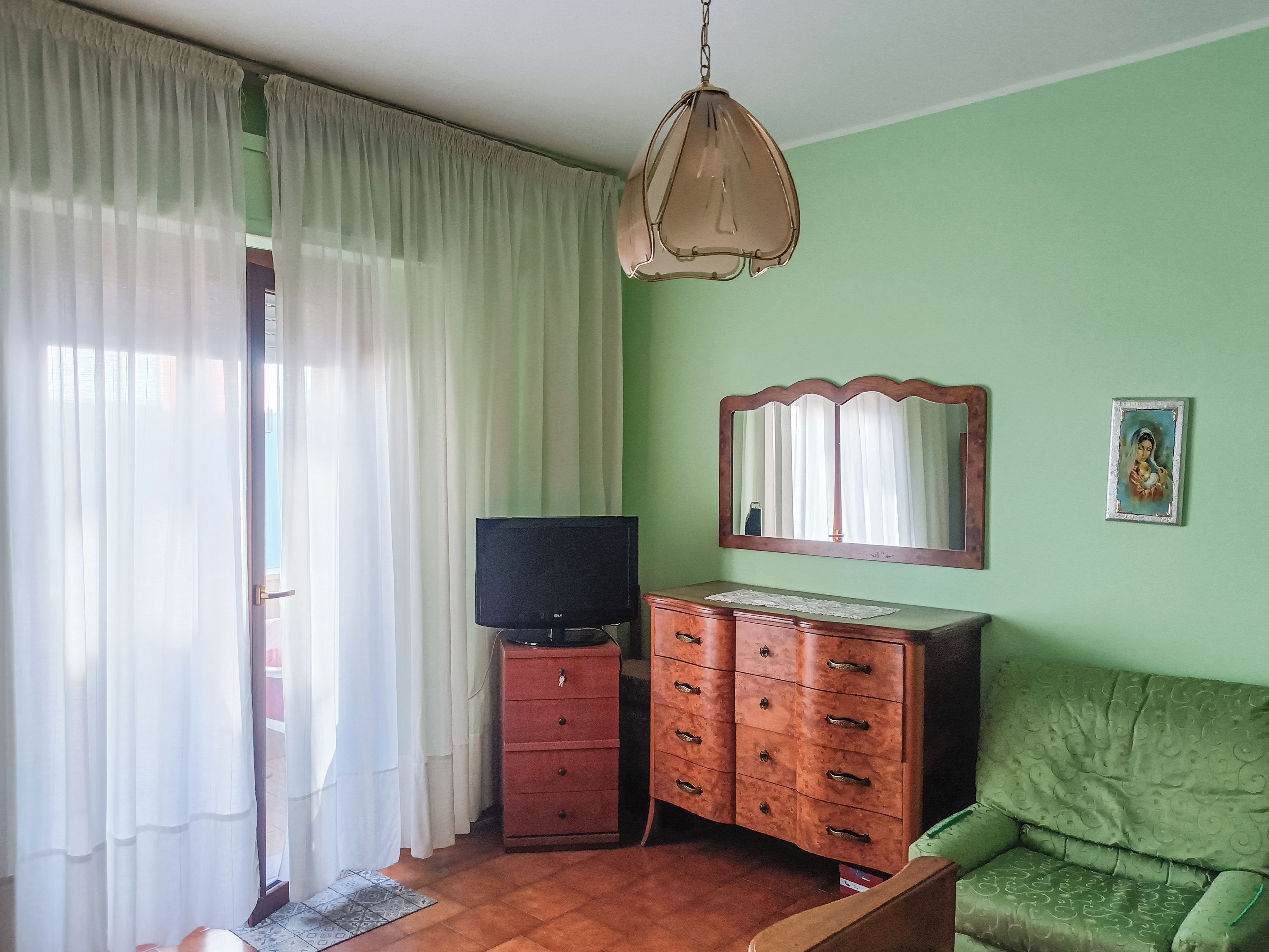 Una habitación con televisión sobre un mueble de madera, un sofá verde, un espejo en la pared y una ventana con cortinas.