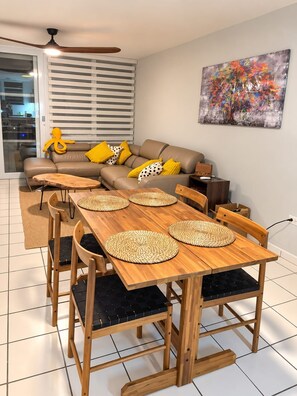 A modern living room with a sofa, a dining table with chairs, a ceiling fan, and a painting on the wall. - Traducción (Ceiba)