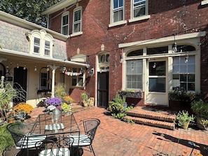 A brick building with a patio area featuring a table and chairs, surrounded by potted plants and flowers. - “2-bedroom apartment in a historic carriage house with garden” (Lambertville)